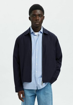 Junger Mann mit einer dunkelblauen Jacke mit Reißverschluss über einem hellblauen Hemd mit Knöpfen und hellblauen Jeans, stehend vor einem schlichten weißen Hintergrund.