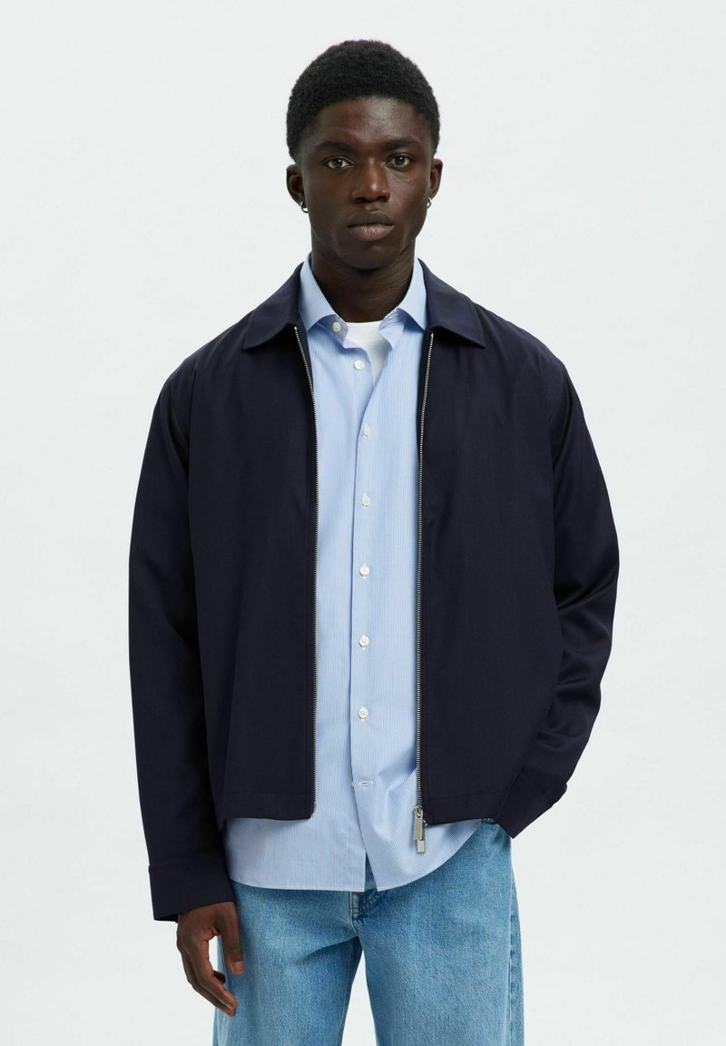 Mladý muž v tmavomodrej bunde na zips cez svetlomodrú košeľu na gombíky a svetlomodré rifle, stojaci pred jednoliatym bielym pozadím.