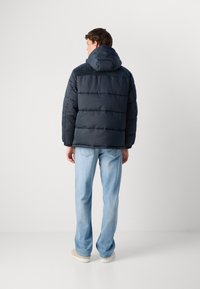 Ναυτικό μπουφάν με κουκούλα, διαθέτει οριζόντια ραφή και υφασμάτινες επιφάνειες στην πάνω πλάτη, φορεμένο με ανοιχτό μπλε τζιν και κρεμ sneakers.