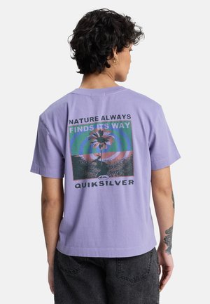 Personne aux cheveux courts et bouclés portant un t-shirt lavande avec une fleur graphique et le texte « La nature trouve toujours son chemin » dans le dos.