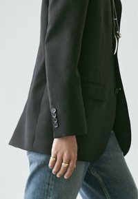 Zwarte, op maat gemaakte blazer met een enkele knoopsluiting, afgeronde revers en zes knopen op de mouwmanchet. Gecombineerd met een blauwe spijkerbroek.