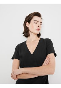 Schwarzes T-Shirt mit kurzem Arm und V-Ausschnitt. Glatte Textur und taillierte Silhouette. Model steht mit verschränkten Armen vor einem schlichten Hintergrund.