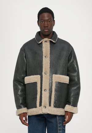 Sort shearling-jakke med beige fleece-trim, knaplukning og to store forlommer. Struktureret læderfinish, krave-design.