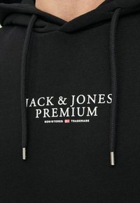 Čierna mikina s kapucňou a šnúrkami s kovovými koncovkami. Na prednej strane sa nachádza logo "JACK & JONES PREMIUM" v bielej farbe. Vyrobená z mäkkého materiálu, má voľný a neformálny dizajn.