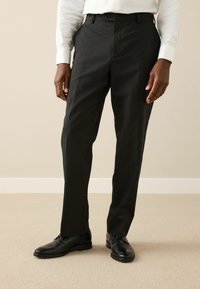 Fekete nadrág egyenes szabással, sima anyagból készült, gombos záródással és övhurkokkal. Fekete cipővel viselve.