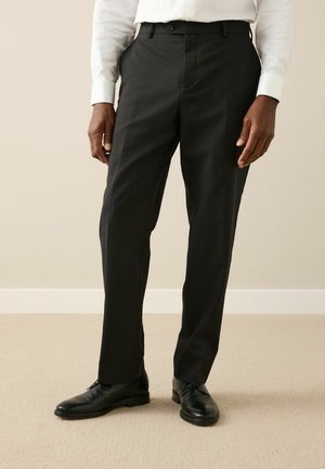 Černé kalhoty s rovnými nohavicemi, vyrobené z hladkého materiálu, s knoflíkovým zapínáním a poutky na opasek. Nosič má na sobě černé boty.