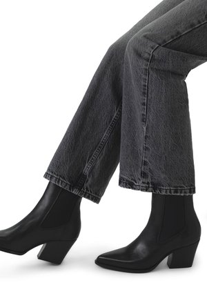 Zwarte leren enkelboots met een spitse neus en een stevige hak, gedragen met een grijze spijkerbroek. De boots hebben zij-elastische panelen voor gemakkelijk aan- en uittrekken.