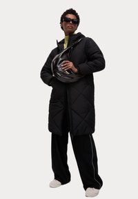 Cappotto imbottito nero trapuntato con cappuccio, indossato sopra una maglietta gialla, abbinato a pantaloni neri oversize e una borsa a tracolla nera lucida.