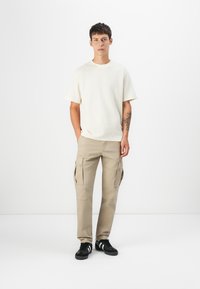 Beige cargo nadrágok több oldalsó zsebbel, laza illeszkedésű krémszínű pólóval párosítva. A modellen fekete sneakers van, fehér részletekkel.
