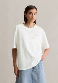 T-shirt en coton blanc surdimensionné avec des manches courtes, présentant un petit détail imprimé sur la poitrine. Assorti à une jupe en denim bleu clair.