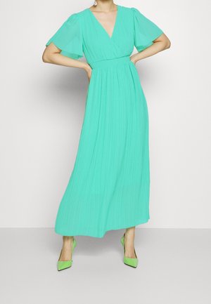 Maxikleid aus leuchtend türkisfarbenem Stoff mit plissiertem Design, V-Ausschnitt und flügelärmel. Kombiniert mit passenden grünen spitzen High Heels.