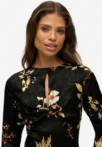Czarna sukienka w kwiaty z długimi rękawami, z dekoltem w kształcie łezki i marszczeniami w talii. Kwiatowe wzory w odcieniach kremowym, żółtym i pomarańczowym.