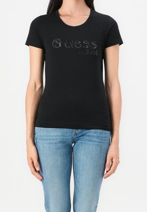 Kvinde iført en sort tætsiddende t-shirt med "Guess est. 1981" i hævede prikker og lyseblå jeans mod en ensfarvet hvid baggrund.