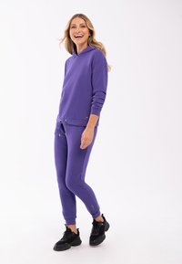 Fioletowy hoodie z kapturem i pasujące dresy wykonane z mieszanki bawełny. Posiadają sznurki, elastyczne mankiety oraz akcent w postaci logo. Noszone z czarnymi sneakersami.