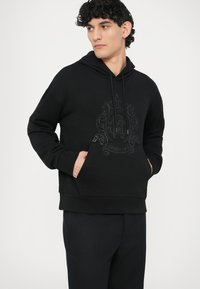 Czarna bluza z kapturem z dużym tonowanym logo na przodzie, posiada kieszeń kangurka i ściągacze przy mankietach, wykonana z miękkiego materiału.