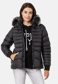 Schwarze Pufferjacke mit abnehmbarer Kunstfellkapuze, gestepptem Textur, Frontzip-Taschen und gerippten Bündchen. Getragen über einem schwarzen Pullover.