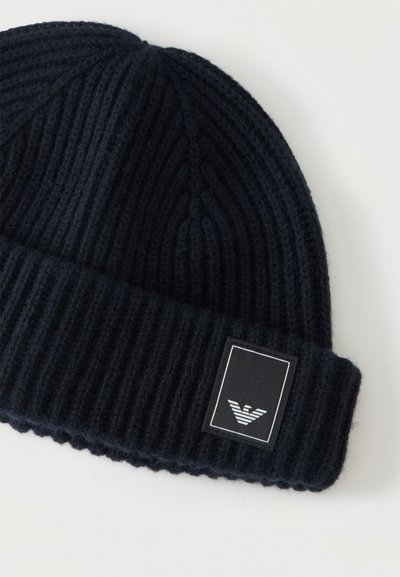 Bonnet en maille côtelée noire avec bord replié présentant un patch logo rectangulaire avec un emblème d'oiseau stylisé en blanc.
