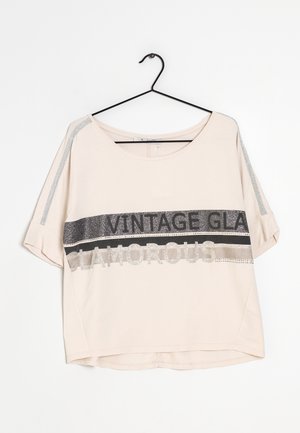 Beige overhemd met korte mouwen met glinsterende zilveren strepen op de mouwen en een tekst op de voorkant die "VINTAGE GLAM GLAMOROUS" luidt, hangend aan een zwarte hanger.