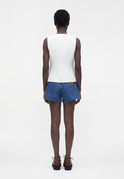 Personne aux cheveux courts portant un haut blanc sans manches, un short en jean bleu et des chaussures à plateforme marron, de dos sur un fond uni.