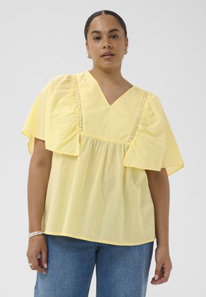 Mujer con una blusa suelta de color amarillo pálido con mangas abullonadas y ribete de encaje, combinada con vaqueros azules, de pie contra un fondo liso.