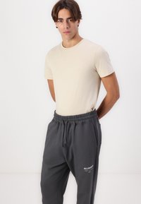 Beige T-Shirt mit kurzen Ärmeln, kombiniert mit dunkelgrauen Jogginghosen, die über einen elastischen Bund und ein Logo-Detail an der Vorderseite verfügen. Weicher, lässiger Stoff.