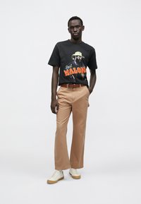 Tricou negru din bumbac cu imprimeu grafic și text, asortat cu pantaloni maro deschis și adidași bej. Modelul stă încrezător, cu mâinile în buzunare.