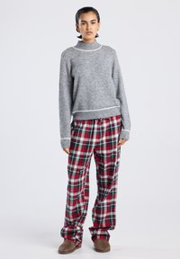 Femme portant un pull gris avec des bords blancs, un pantalon écossais rouge et vert, et des pantoufles marron, se tenant devant un fond blanc uni.