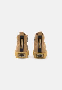 Bottines en nubuck beige avec une semelle texturée jaune, dotées de poignées en tissu noir et d'une fermeture éclair sur le côté pour un accès facile.