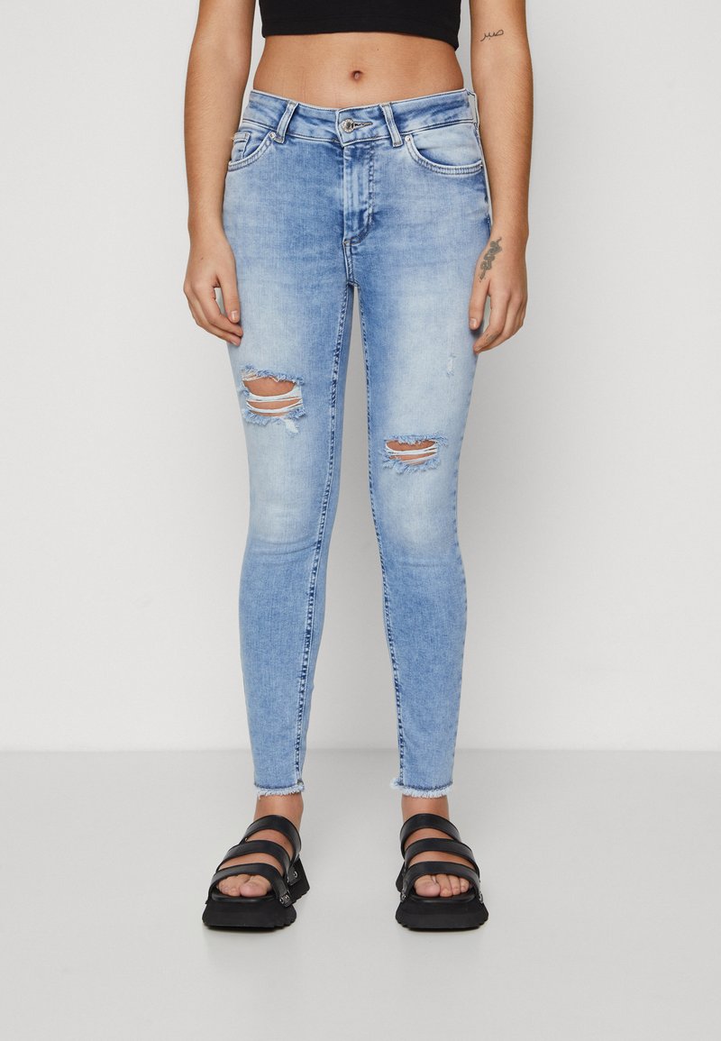 Light blue skinny jeans with ripped accents at the knees, finished with a frayed hem. Paired with black strappy sandals.