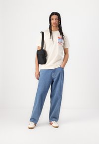 Beige cotton T-shirt with graphic print, loose-fitting blue denim pants, and white sneakers with red stripes. Black crossbody bag on shoulder.