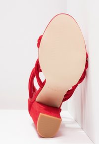 Escarpin en daim rouge avec une semelle épaisse, des sangles croisées et une semelle beige lisse. Présente un léger style plateforme.