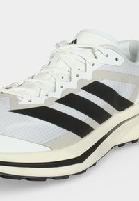 Άσπρο και γκρι αθλητικό παπούτσι τρεξίματος με μαύρο λογότυπο τριών ραβδώσεων, λευκά κορδόνια και μαξιλαρωμένη σόλα σχεδιασμένο για αθλητική χρήση.