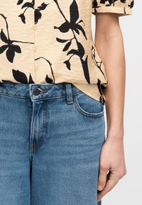 Beige overhemd met zwarte bloemenprint, ingestopt in blauwe spijkerbroek; rechterarm ontspannen langs het lichaam met hand gedeeltelijk zichtbaar.