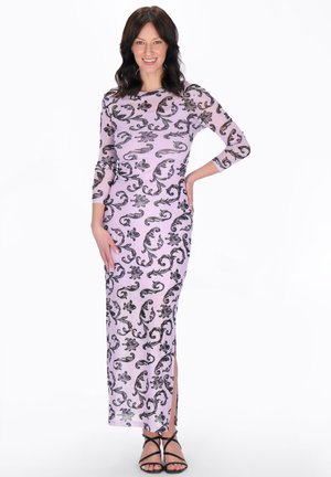 Nő áll, egyik kezével a csípőjén, hosszú levendulaszínű ruhát visel fekete virágmintákkal és fekete pántos magassarkúval, mosolyogva a kamerába.