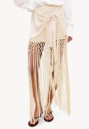 Beige wikkelrok met grote strik en lange macramé franjes die de benen bedekken, gecombineerd met een wit overhemd en zwarte sandalen aan de voeten.