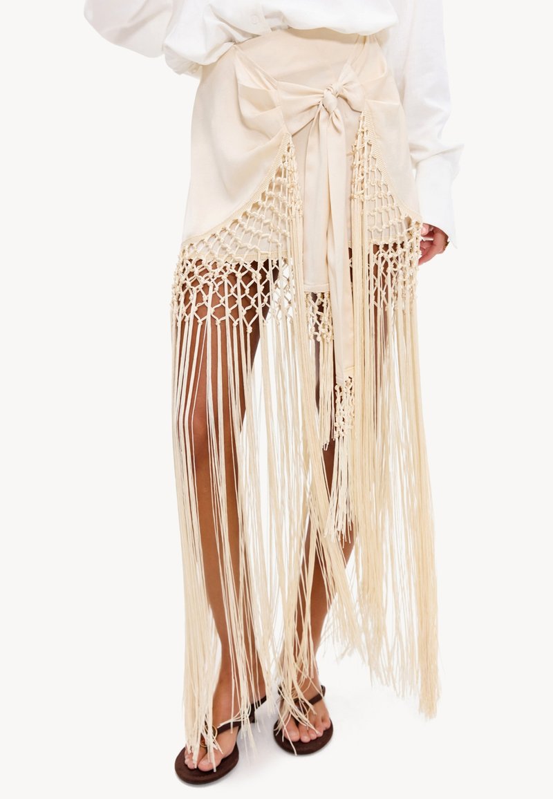Jupe portefeuille beige avec grand nœud et longues franges en macramé couvrant les jambes, associée à une chemise blanche et des sandales noires aux pieds.