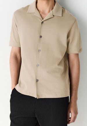 Homme portant une chemise beige à manches courtes boutonnée avec col et un pantalon noir texturé, debout les mains dans les poches.