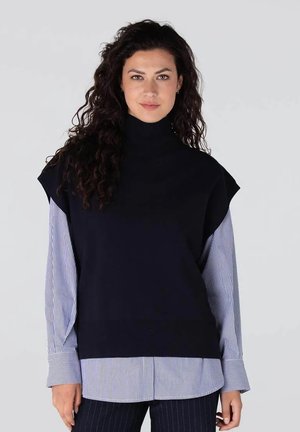 Marineblauwe oversized gebreide turtleneck trui, gelaagd over een lichtblauw en wit gestreept shirt met brede mouwen. Gladde textuur en relaxte pasvorm.