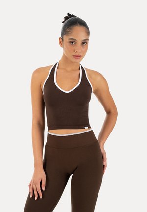 Femme aux cheveux attachés en chignon portant un crop top marron à col halter avec une bordure blanche et des leggings taille haute assortis sur un fond uni.