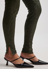 Olivgröna paljettbyxor med vertikal ribbning, med sidslitsar vid anklarna. Matcherade med svarta mules med spetsig tå och tunna remmar.