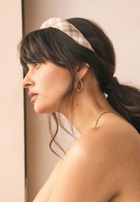 Beige-sävyinen ruudullinen hiuspanta, kultaiset renkaankorvakorut ja teksturoitu, paljas olkapää. Hiukset tyylitelty löysiin laineisiin. Neutraali tausta.