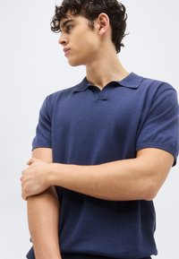 Νεαρός άνδρας με καστανά σγουρά μαλλιά, φοράει μπλε σκούρο κοντομάνικο πόλο, κοιτάει αριστερά με σταυρωμένα τα χέρια στο στήθος.