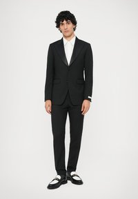 Muž s kučeravými vlasmi stojí, oblečený v čiernom obleku so saténovými klopami, bielej košeli a čierno-bielych mokasínach s detailmi zlatého reťazca.