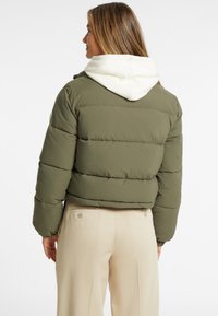 Tamsiai žalia pūkinė striukė su balta kapuce, kuri turi siuvinėjimo detales ir trumpą dizainą, derinama su smėlio spalvos kelnėmis.