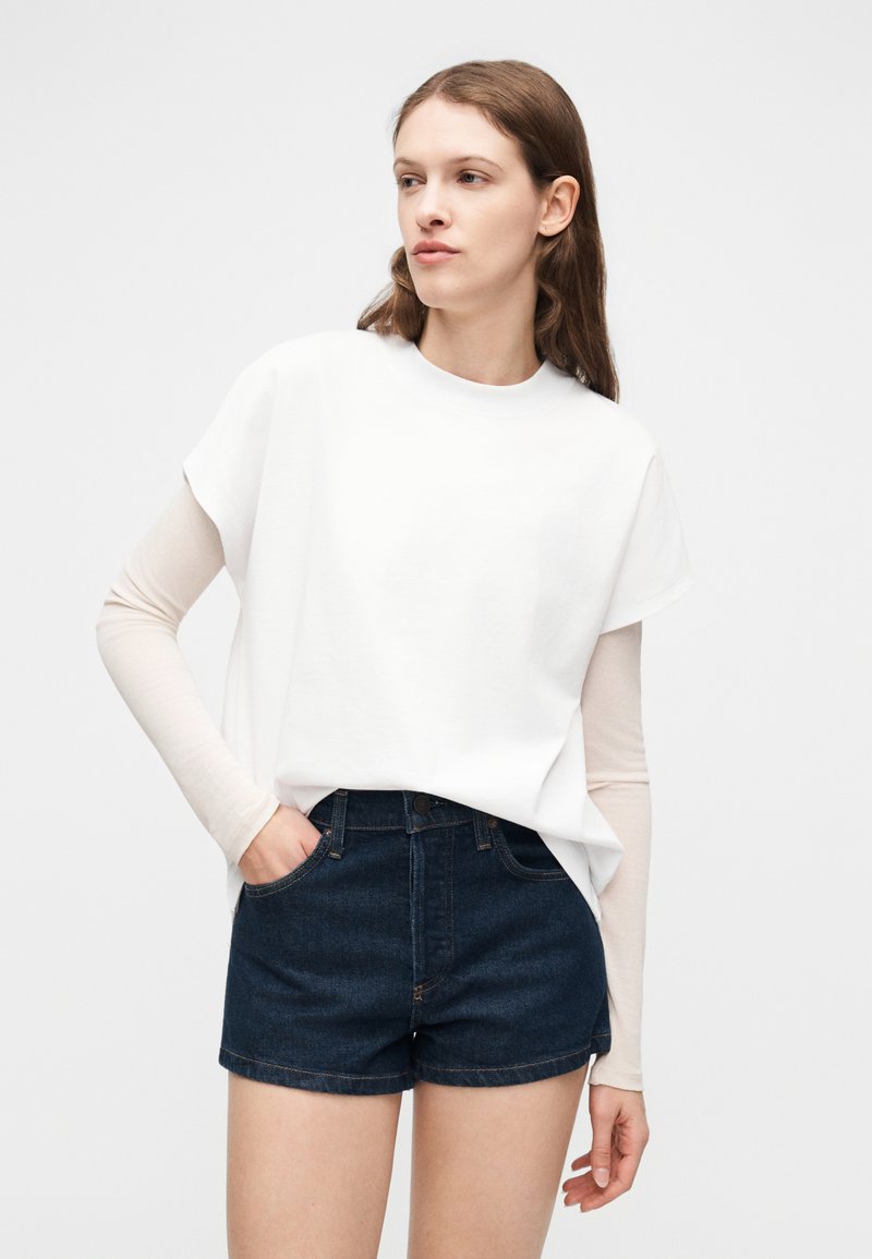 Femme aux cheveux bruns portant un haut blanc à manches longues superposé avec un débardeur blanc, short en jean foncé, debout avec une main dans la poche.