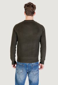 Uomo con capelli castani corti che indossa un maglione a coste verde scuro e jeans chiari sbiaditi, visto da dietro su uno sfondo neutro.