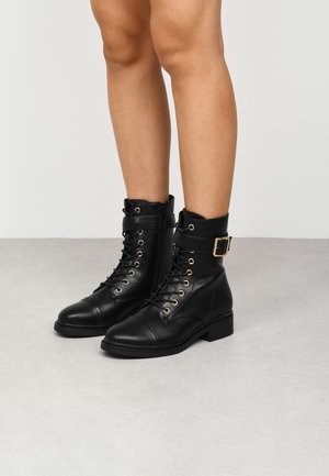 Bottines en cuir noir avec des œillets dorés, des lacets, une fermeture éclair sur le côté et une sangle à boucle ; bout rond et petit talon épais.