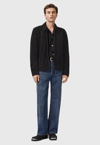 Mand står iført en sort ruskindsjakke, sort skjorte, blå jeans, sort bælte og sorte sko med metalnæser.