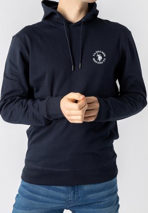 Person trägt dunkelblaue U.S. Polo Assn. Kapuzenjacke mit Kordelzug und blaue Jeans, Hände vor dem Körper verschränkt, vor einfarbig hellem Hintergrund.