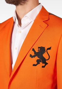 Pomarańczowy żakiet z czarnym haftem lwa. Posiada wyraźnie zarysowane ramiona, klapki i gładką teksturę. Pod spodem biała koszula.